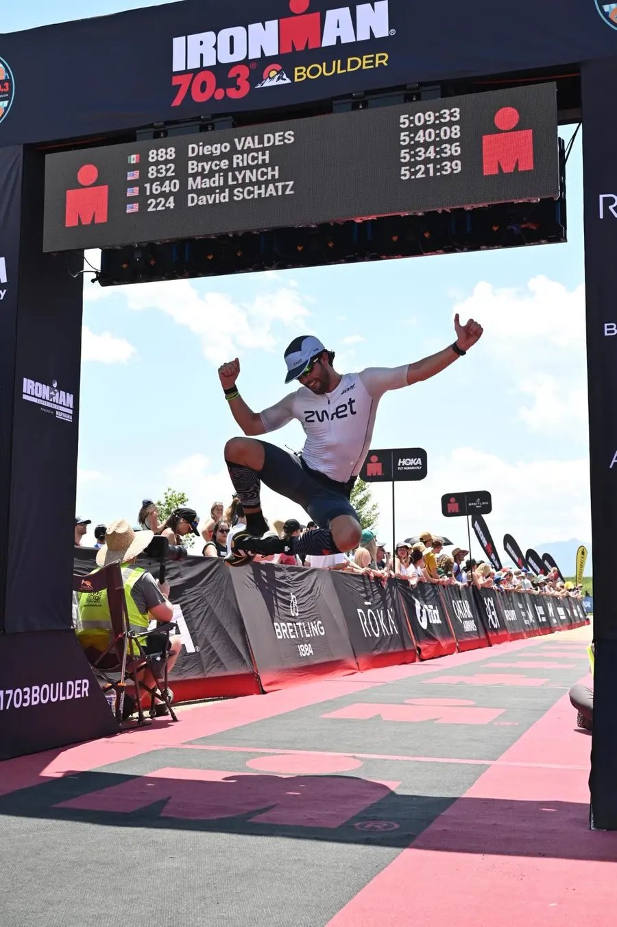 Diego jumping through the Ironman 70.3 Boulder finish arch, his name and 5:09:33 on the board above.